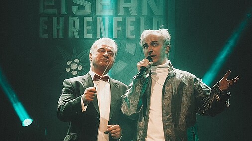 Foto zum Thema "Claus Weselsky und Jan Böhmermann": Zu sehen sind Weselsky und Böhmermann auf der Bühne der Jahrhunderthalle in Frankfurt. Weselsky hat einen Taktstock in der Hand, Böhmermann ein Mikrofon.