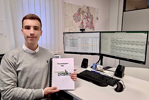 Foto zum Thema "wie wird man Kämmerer": Zu sehen ist Niklas Riedinger, Kämmerer der Gemeinde Forbach. Er sitzt am Schreibtisch und hält den Haushaltsplan in der Hand.